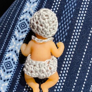May include: A small doll with a light skin tone, wearing a crocheted gray hat and diaper. The doll is lying on its stomach, with its arms and legs slightly extended. The background is a blue fabric with a white geometric pattern.