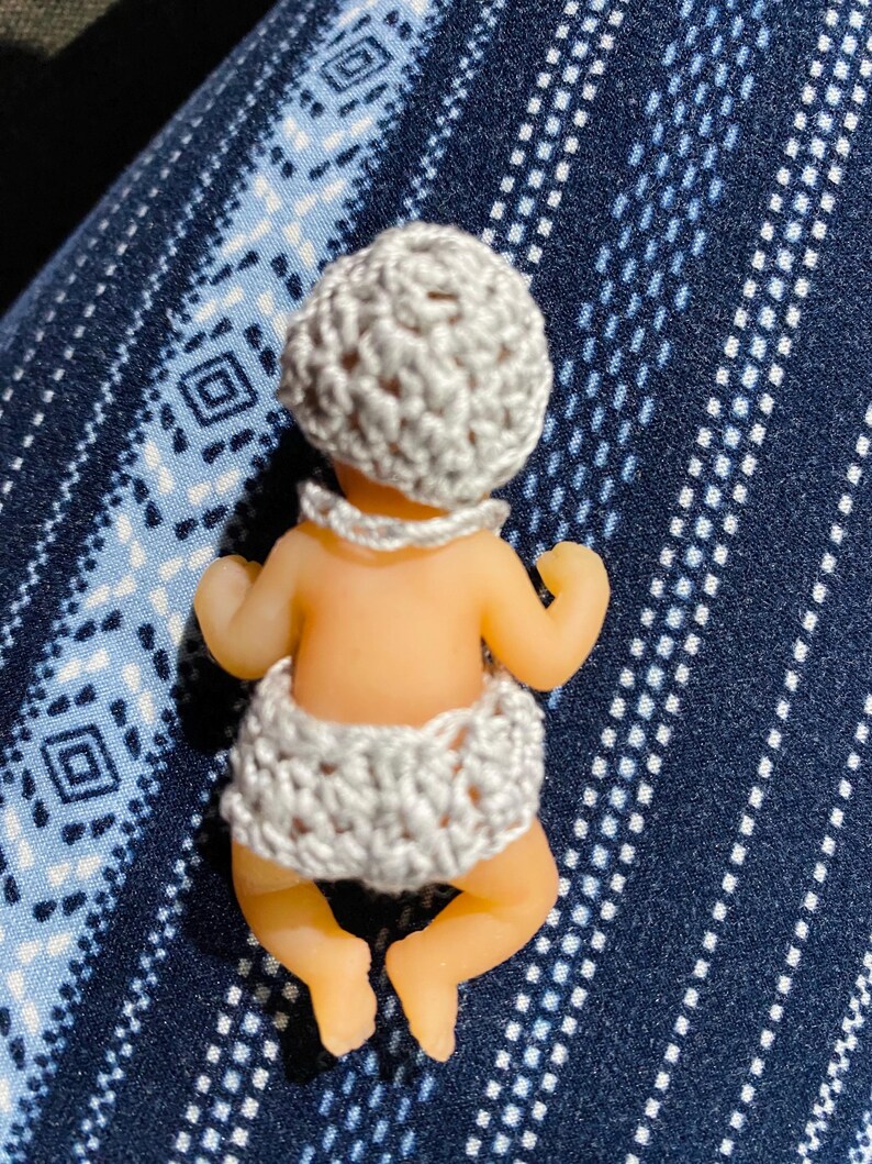 May include: A small doll with a light skin tone, wearing a crocheted gray hat and diaper. The doll is lying on its stomach, with its arms and legs slightly extended. The background is a blue fabric with a white geometric pattern.