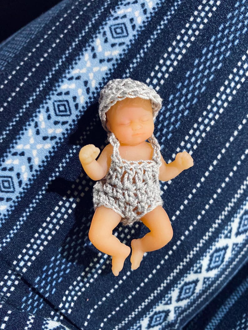 May include: A miniature baby doll with a gray crocheted outfit and matching hat. The doll has a realistic skin tone and is lying down. The background is a dark blue fabric with a white geometric pattern.