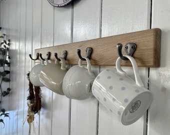 May include: A wooden wall-mounted mug rack with five hooks. The rack is made of natural wood and has a rustic finish. Four white mugs with different patterns are hanging on the hooks.