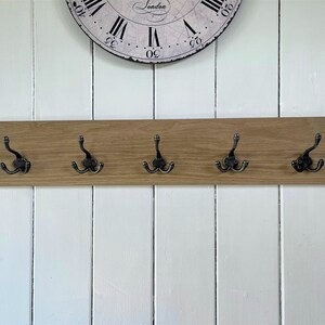 May include: A wooden wall-mounted coat rack with five hooks. The rack is made of light brown wood and has a natural finish. The hooks are black metal and have a distressed finish.