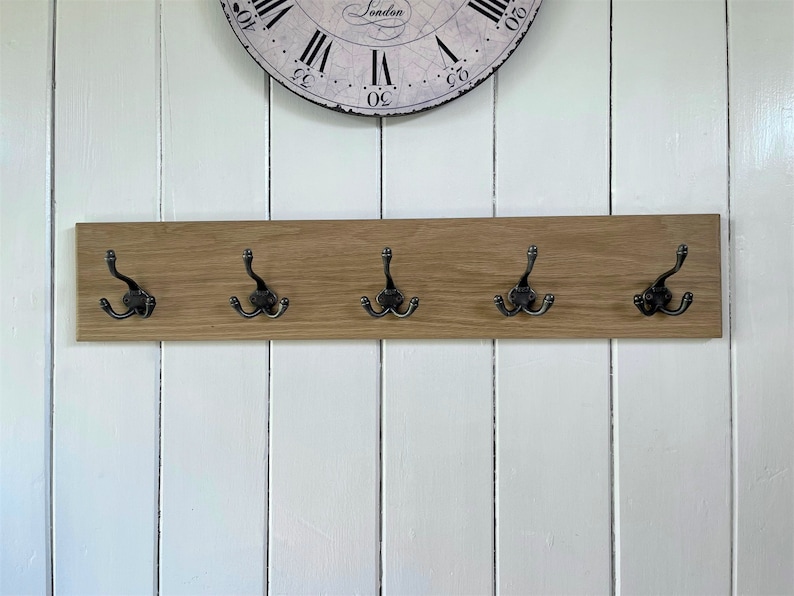 May include: A wooden wall-mounted coat rack with five hooks. The rack is made of light brown wood and has a natural finish. The hooks are black metal and have a distressed finish.