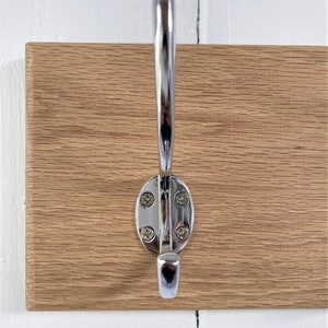 May include: A close-up of a chrome hook mounted on a wooden wall-mounted coat rack. The hook has a rounded oval base and a single hook.