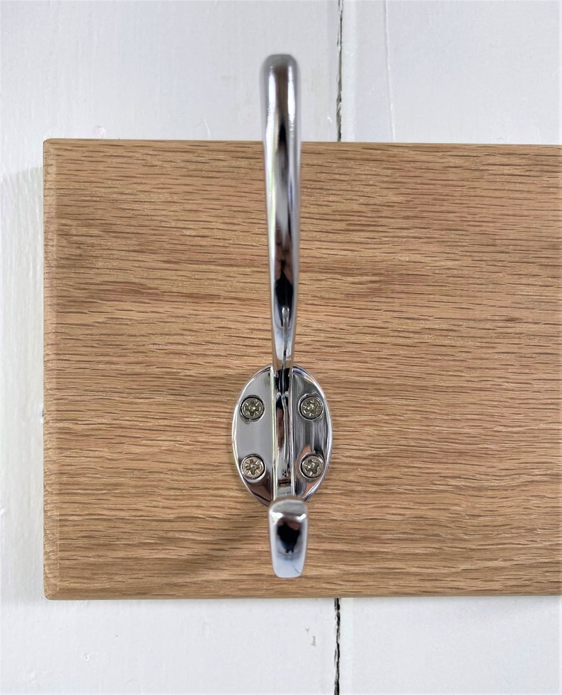 May include: A close-up of a chrome hook mounted on a wooden wall-mounted coat rack. The hook has a rounded oval base and a single hook.
