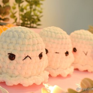 May include: Three handmade, white crochet ghost plushies with different facial expressions. Each ghost has black button eyes and a small mouth. The plushies are arranged on a pink surface with string lights and greenery in the background.