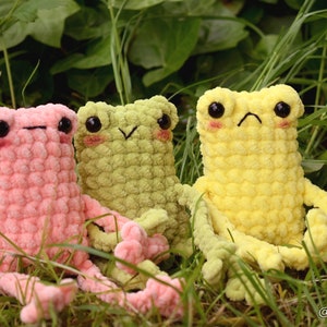 May include: Three crocheted frog plushies in pink, green, and yellow. The frogs are sitting in a bed of green grass.