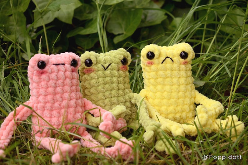 May include: Three crocheted frog plushies in pink, green, and yellow. The frogs are sitting in a bed of green grass.