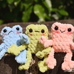 May include: Three crocheted frog plushies sitting on a wooden surface. The frogs are light blue, light green, and light pink. Each frog has black eyes and a small smile.