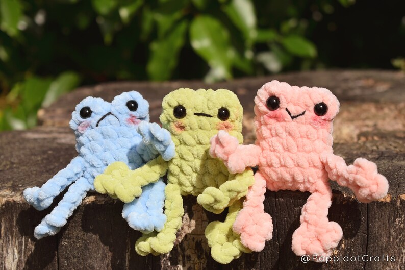 May include: Three crocheted frog plushies sitting on a wooden surface. The frogs are light blue, light green, and light pink. Each frog has black eyes and a small smile.
