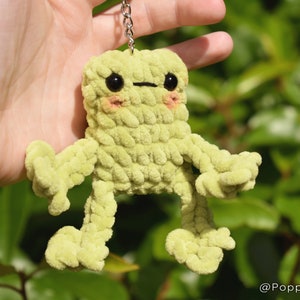 May include: A green crocheted frog keychain with black eyes and pink cheeks. The frog has long, thin arms and legs.