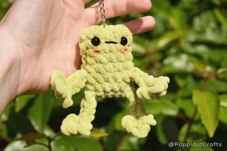 May include: A green crocheted frog keychain with black eyes and pink cheeks. The frog has long, thin arms and legs.