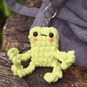 May include: A lime green crocheted frog keychain with black eyes and pink cheeks. The frog is sitting on a wooden surface.