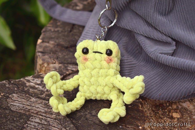 May include: A lime green crocheted frog keychain with black eyes and pink cheeks. The frog is sitting on a wooden surface.