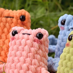 May include: Four crocheted plush frog toys in different colors: orange, pink, blue, and green. The frogs have black eyes and are sitting on a bed of green grass.