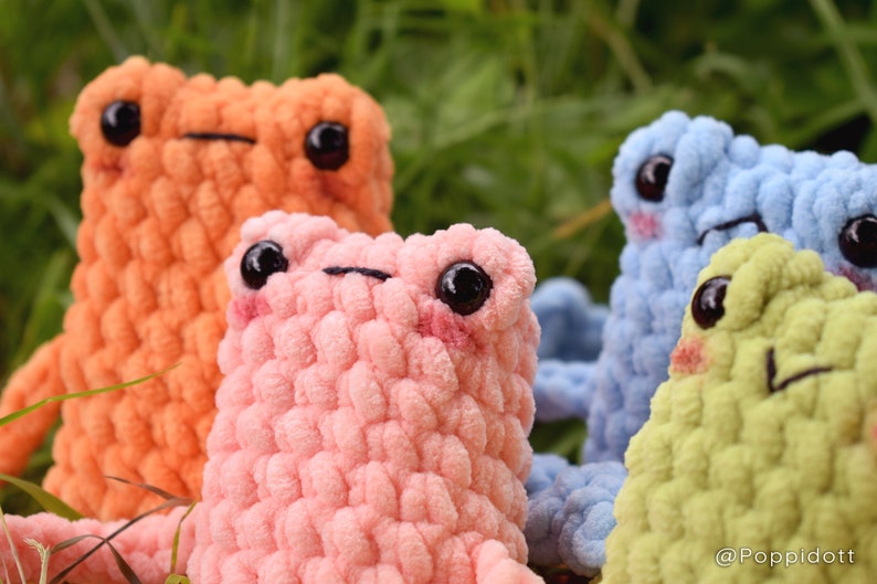 May include: Four crocheted plush frog toys in different colors: orange, pink, blue, and green. The frogs have black eyes and are sitting on a bed of green grass.