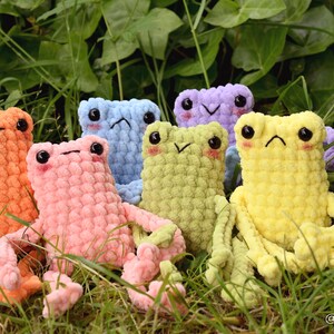 May include: A group of seven crocheted frog plushies in various pastel colors, including orange, pink, light blue, green, yellow, and purple. The plushies are sitting in a bed of green grass.
