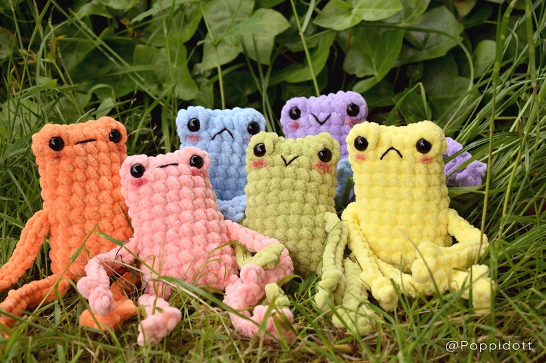 May include: A group of seven crocheted frog plushies in various pastel colors, including orange, pink, light blue, green, yellow, and purple. The plushies are sitting in a bed of green grass.