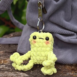 May include: A lime green crocheted frog keychain with black eyes and pink cheeks. The frog is sitting on a wooden surface and is attached to a silver keychain.