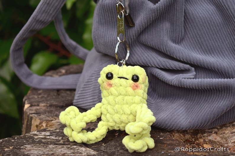 May include: A lime green crocheted frog keychain with black eyes and pink cheeks. The frog is sitting on a wooden surface and is attached to a silver keychain.