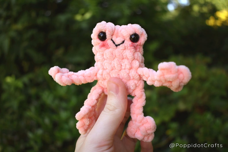 May include: A pink crocheted frog plush toy with black eyes and a smiling mouth. The frog is being held by a hand.