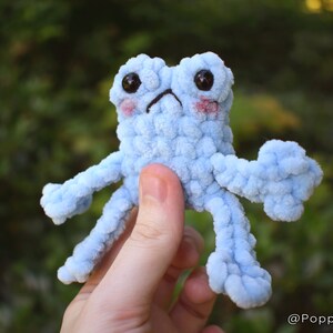 May include: A light blue crocheted frog with large black eyes and pink cheeks. The frog has four long, thin arms and legs.