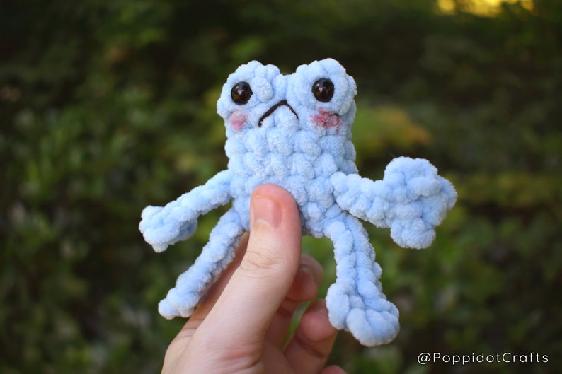 May include: A light blue crocheted frog with large black eyes and pink cheeks. The frog has four long, thin arms and legs.
