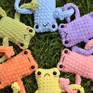 May include: Five crocheted frog plushies in different colors, including green, orange, pink, purple, and yellow, are arranged in a circle on a bed of green grass. The frogs have black eyes and are all looking up at the camera.