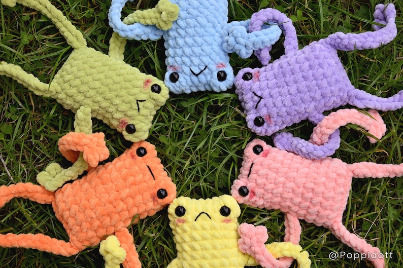 May include: Five crocheted frog plushies in different colors, including green, orange, pink, purple, and yellow, are arranged in a circle on a bed of green grass. The frogs have black eyes and are all looking up at the camera.