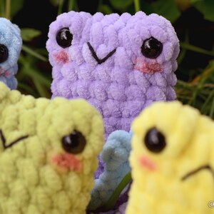 May include: Four crocheted plush toys with different colored fur. The toys have large eyes, blushed cheeks, and embroidered mouths. The toys are arranged in a group, with the purple toy in the center.