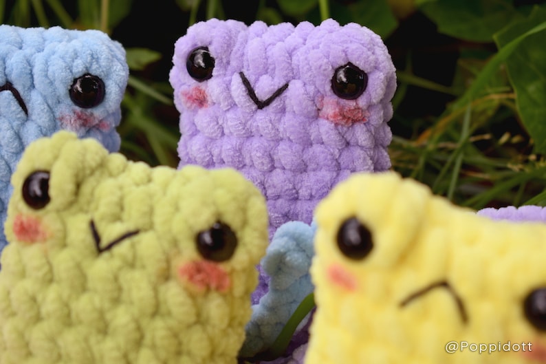 May include: Four crocheted plush toys with different colored fur. The toys have large eyes, blushed cheeks, and embroidered mouths. The toys are arranged in a group, with the purple toy in the center.