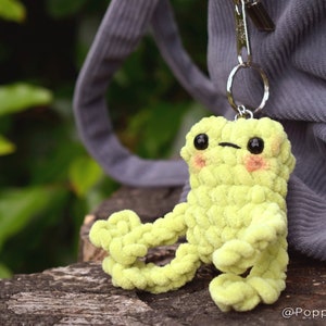 May include: A lime green crocheted frog keychain with black eyes and pink cheeks. The frog is attached to a silver key ring.