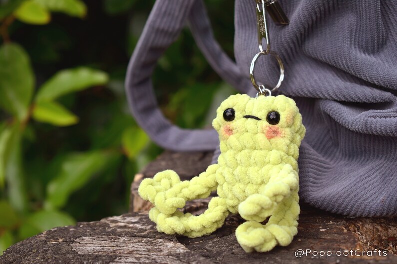 May include: A lime green crocheted frog keychain with black eyes and pink cheeks. The frog is attached to a silver key ring.