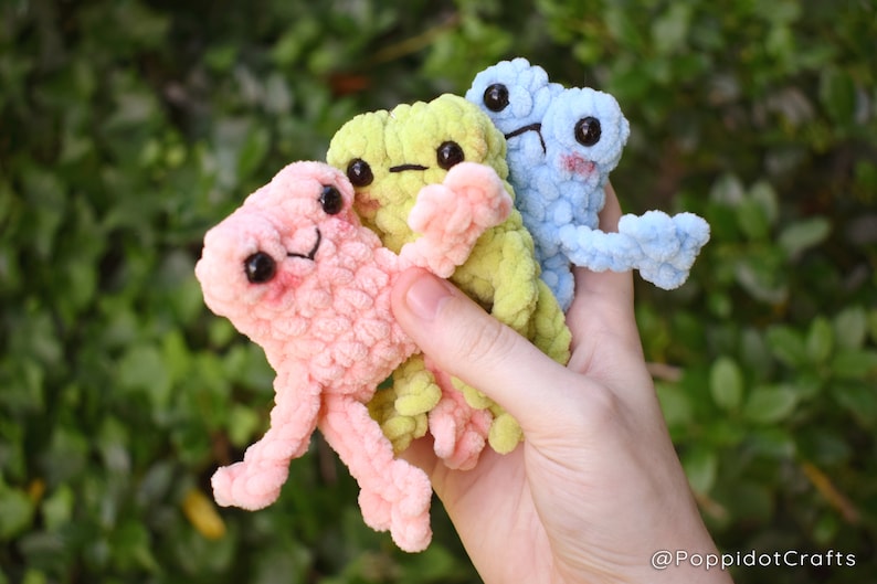 May include: Three small crocheted plush toys shaped like frogs in pink, green, and blue colors. The toys have large black eyes and are held in a hand.
