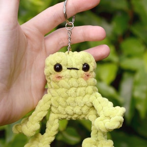 May include: A lime green crocheted frog keychain with black eyes and a small black mouth. The frog has four arms and four legs, all crocheted in the same lime green yarn. The keychain is attached to a silver chain and ring.