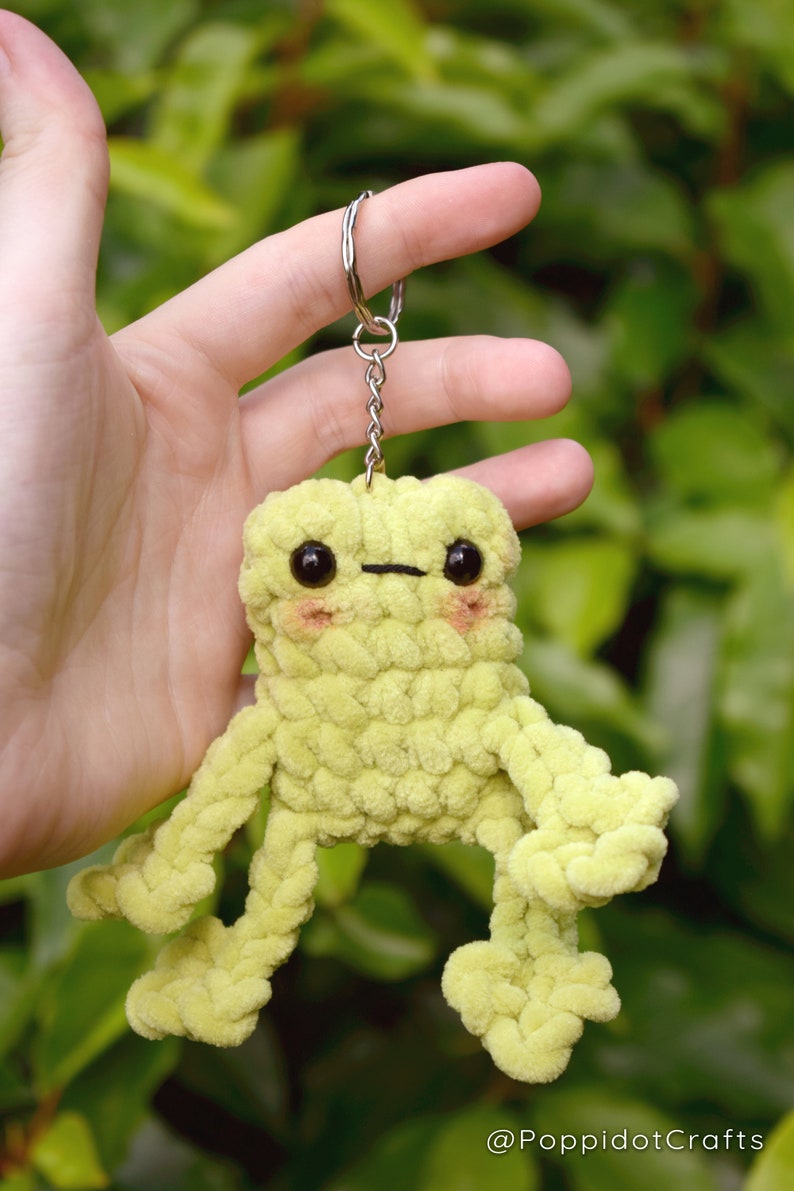 May include: A lime green crocheted frog keychain with black eyes and a small black mouth. The frog has four arms and four legs, all crocheted in the same lime green yarn. The keychain is attached to a silver chain and ring.