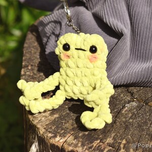 May include: A lime green crocheted frog keychain with black eyes and pink cheeks. The frog is sitting on a tree stump.