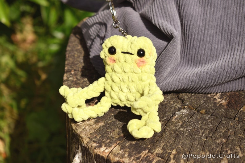 May include: A lime green crocheted frog keychain with black eyes and pink cheeks. The frog is sitting on a tree stump.