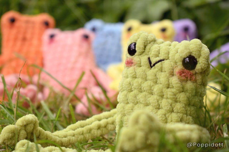 May include: A group of crocheted frog plushies in various colors, including green, pink, orange, blue, and yellow. The frogs are sitting in a bed of green grass.