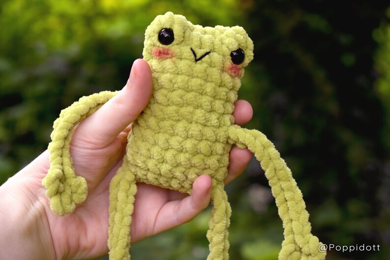 May include: A green crocheted frog plush toy with black eyes and pink cheeks. The frog has long arms and legs.