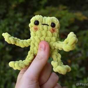 May include: A green crocheted frog toy with black eyes and a small, stitched-on mouth. The frog is being held by a hand.