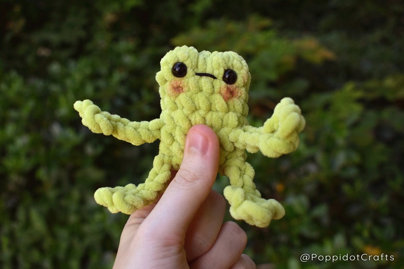May include: A green crocheted frog toy with black eyes and a small, stitched-on mouth. The frog is being held by a hand.