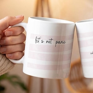 May include: Two white ceramic mugs featuring horizontal light pink stripes and black cursive text. The left mug displays the words 'let's not panic' and the right mug displays the words 'until after coffee'. Both mugs have a smooth, glossy finish.