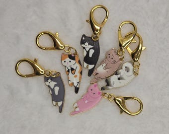 May include: A collection of cat-themed charms with gold-colored clasps. The charms feature various cat designs in colors like black, white, pink, gray, and calico. The cats are in different poses, some standing and others lying down.