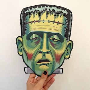 Puede incluir: Máscara de papel verde y amarilla de Frankenstein con la frente cosida y un tornillo de cuello negro y blanco.