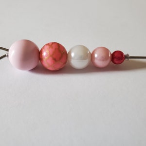 Könnte beinhalten: Ein rosa, weißer und roter Perlenanhänger mit einer silbernen Schlaufe. Die Perlen sind rund und haben eine glatte Oberfläche. Der Anhänger ist an einem Silberdraht befestigt.
