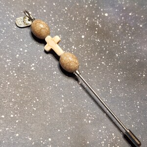 May include: A silver pin with a white cross charm and two brown and white patterned beads. The pin has a silver tag that reads "Made with Love".