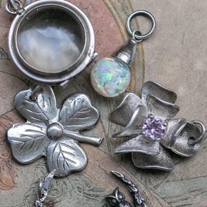 May include: A collection of silver-toned pendants and charms. Includes a locket, a pendant with an opal-like orb, a four-leaf clover, a flower with a purple center, and a crescent moon with three owls. The items are displayed on a patterned surface.