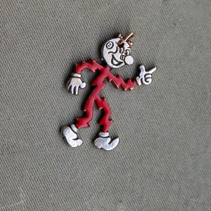 May include: A whimsical pin featuring a cartoon figure with a white face, red body, and silver accents. The figure is pointing with one hand and has a cheerful expression. The pin is set against a textured, olive-green background.
