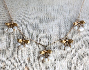 May include: A gold chain necklace with five pearl and gold flower charms. The necklace is on a white fabric background.