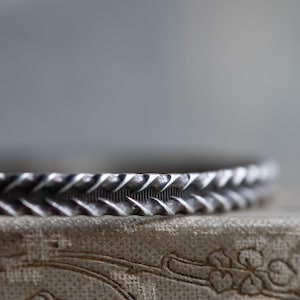 May include: Close-up of a silver-toned bracelet with a textured, repeating pattern. The bracelet's design features a series of pointed, triangular shapes. The background is a soft, neutral color, highlighting the bracelet's intricate details.